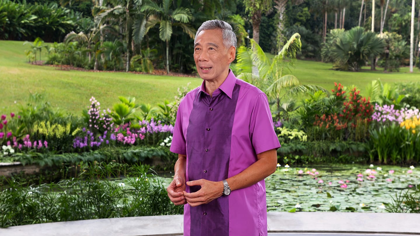 Lee Hsien Loong in a purple shirt, stands in a garden with palms and water lilies.
